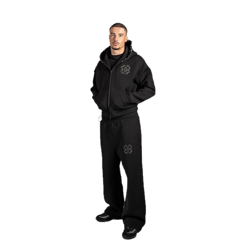 Man in a black zip front hoodie with a clover motif on the chest and black trousers on a white background