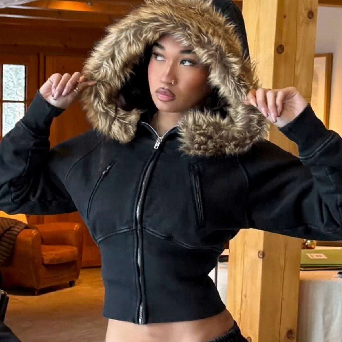Model in a chalet wearing the black cropped fur trimmed hooded zip jacket with the plush hood raised