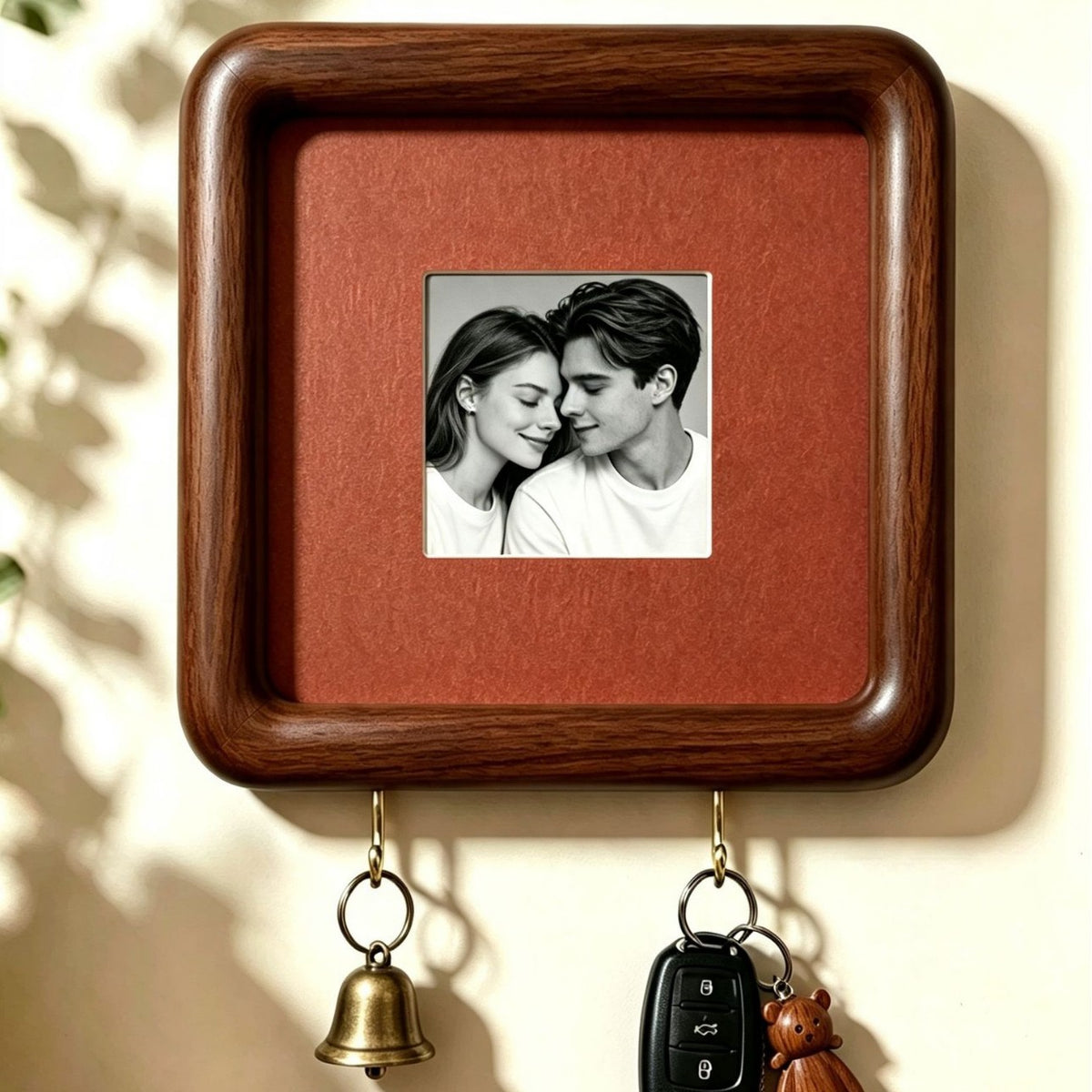Close view of a square dark wood wall frame with plain terracotta backing, a bell and a key fob on two hooks