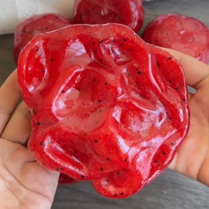 Red watermelon squishy ball pressed deep into a palm, black seeds and pale ice flecks visible
