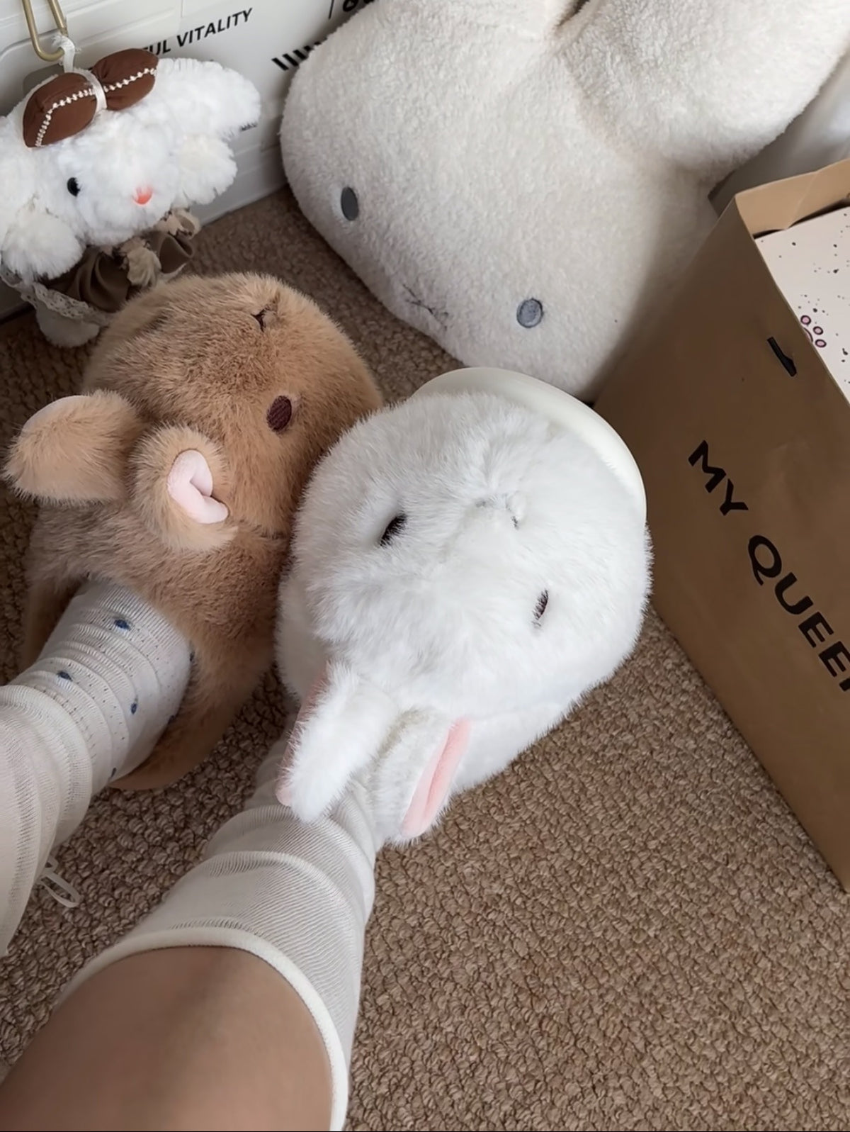 One brown and one white bunny plush slipper worn on feet in light socks on a brown carpet, view 3 of 3