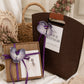 Monet Purple option, wooden gift box with a purple ribbon, orchid flower and card beside a brown bag with a cut-out handle