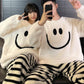 Couple posing in matching white plush smiley face pajama tops and striped trousers, one holding a phone