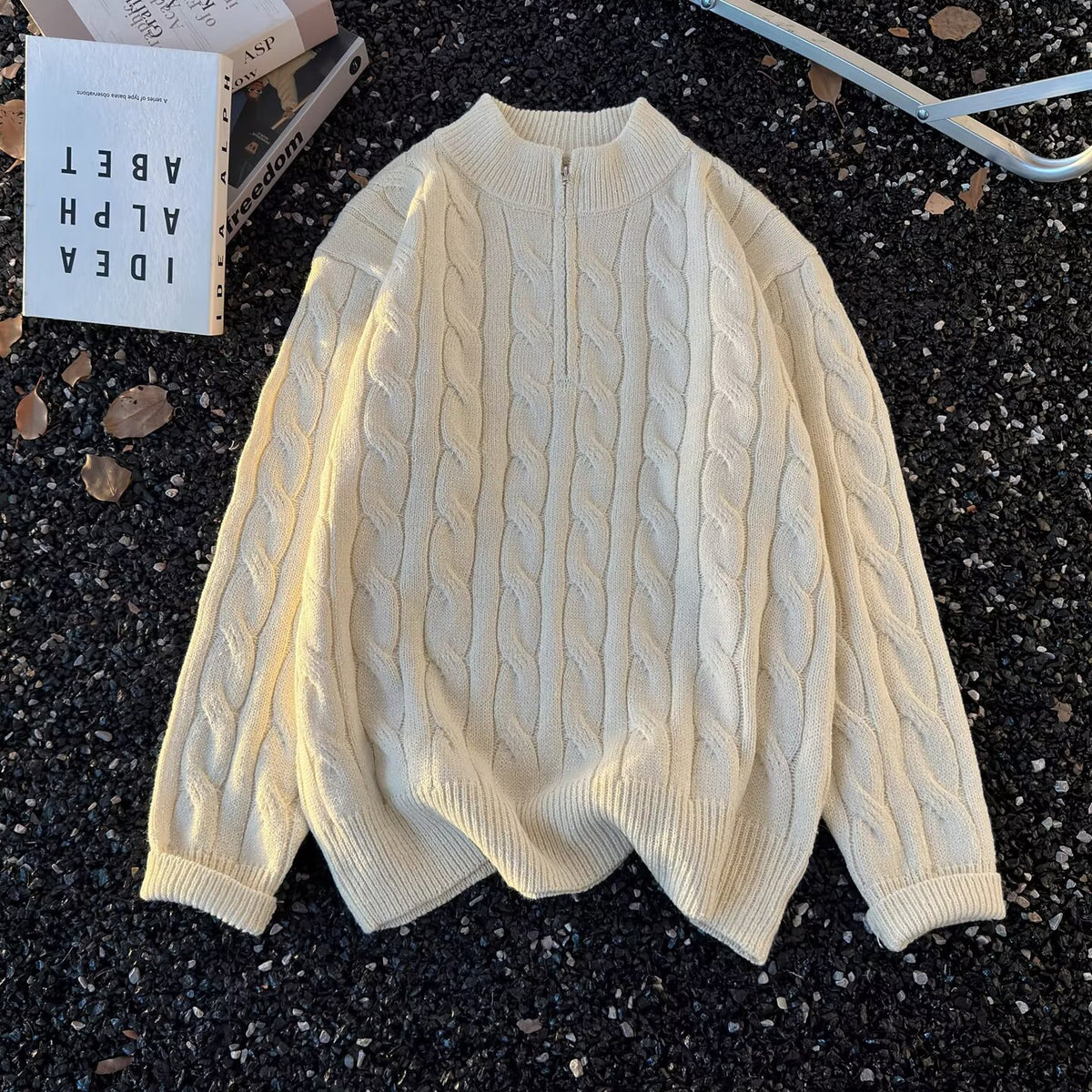 Cream half zip cable knit sweater laid flat on dark gravel beside a book, first cream option photo