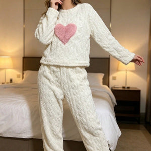 Woman in a cream plush fleece pajama set with a pink heart patch standing beside a bed with warm lamps