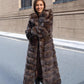 Woman in a long brown vegan fur coat held closed in front, hood down, standing on a street
