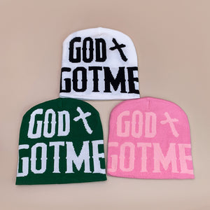 White, green and pink knit beanies with bold GOD cross and GOTME lettering on a beige background
