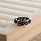 Wide band ring with blackened scrollwork pattern standing on the edge of a wooden ring tray, lettering visible inside