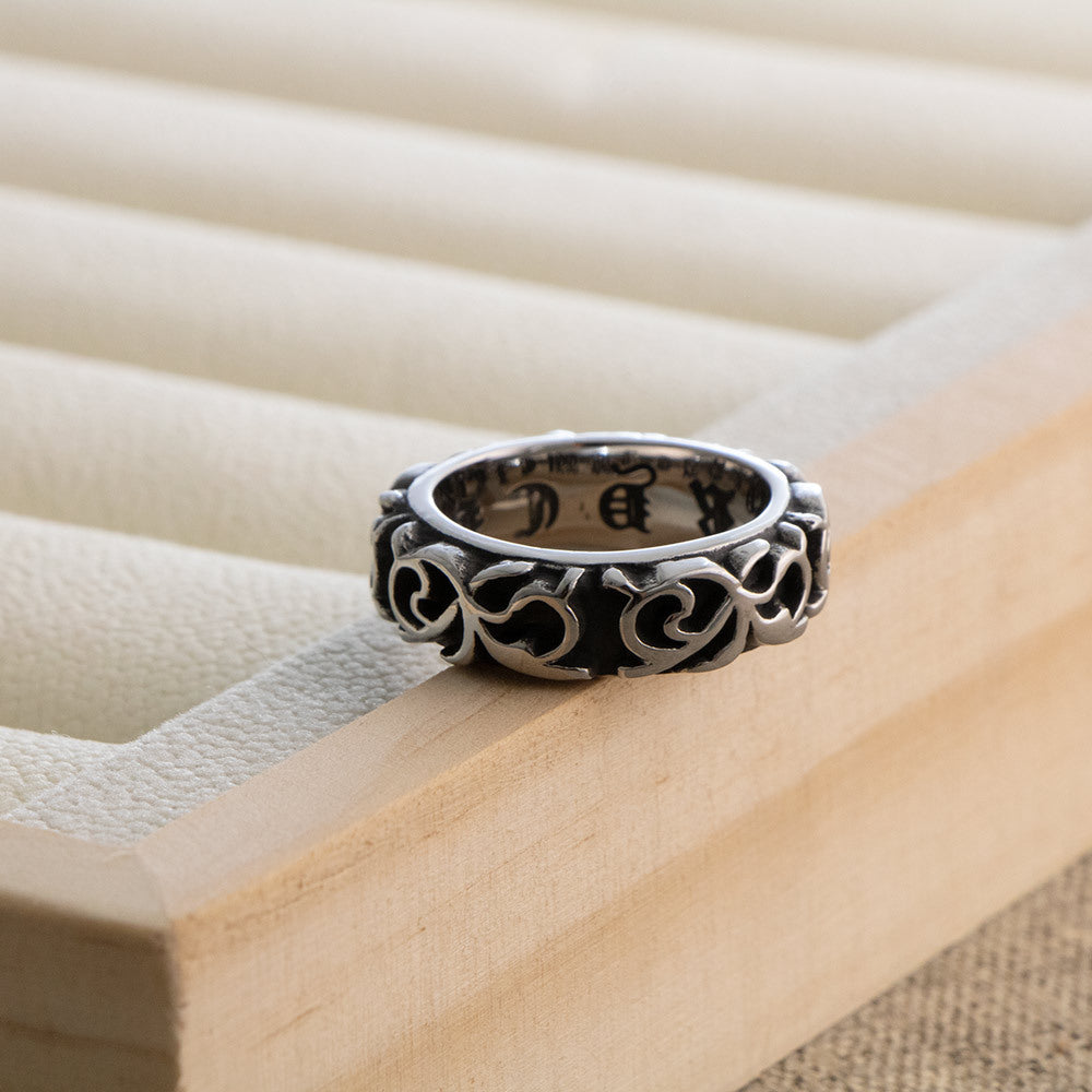 Wide band ring with blackened scrollwork pattern standing on the edge of a wooden ring tray, lettering visible inside