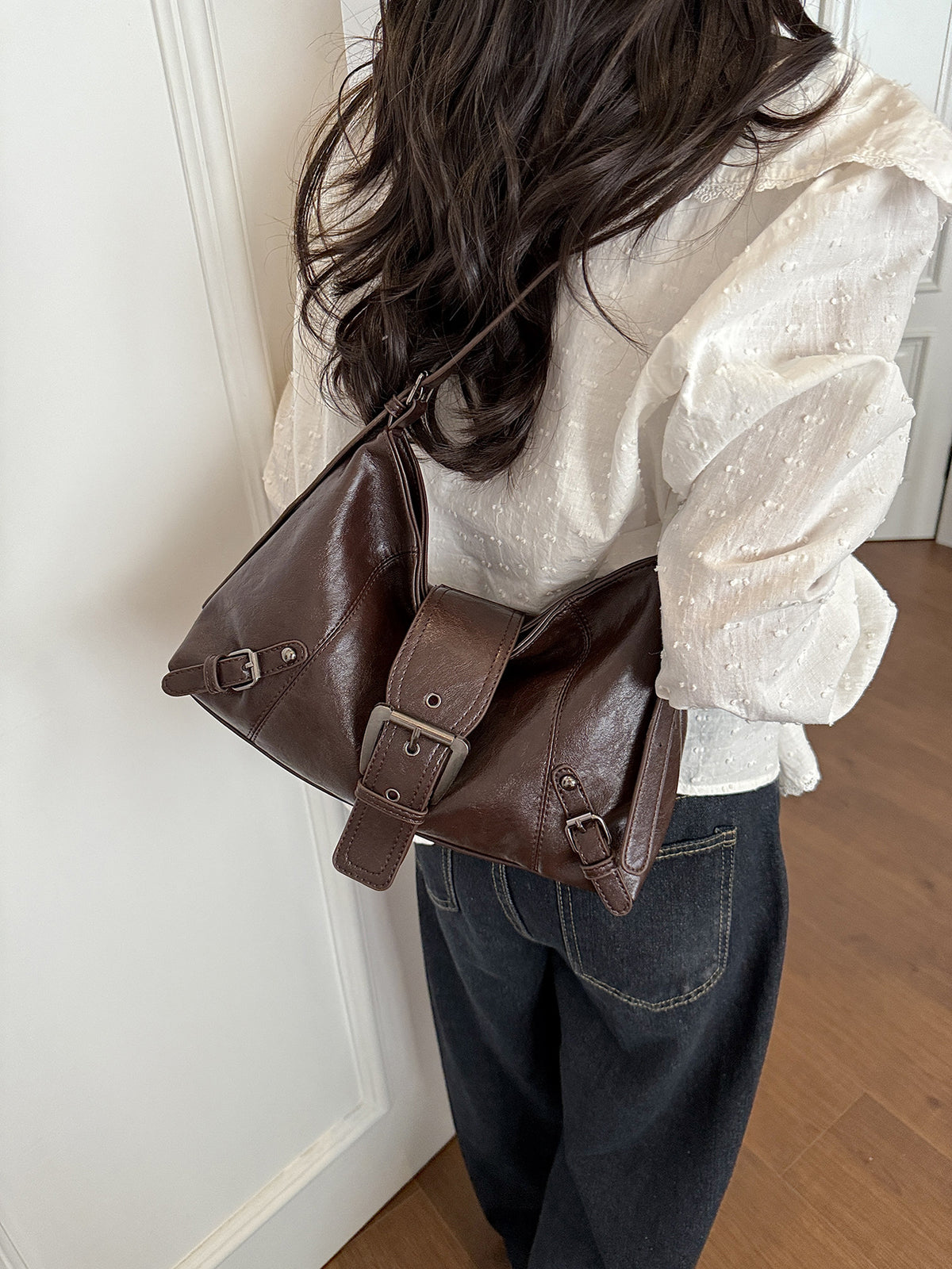Brown soft vegan leather hobo bag with front buckle strap worn on the shoulder, seen from behind with a white blouse