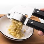 Stainless steel garlic press crushing minced garlic into a white bowl on a wooden board