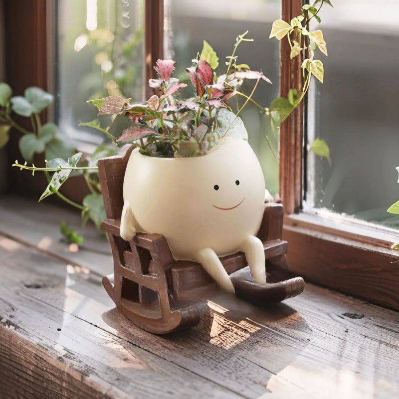Cream smiling face resin planter in a brown rocking chair with a small plant on a windowsill
