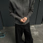 Man in the gray zip knit cardigan with hands clasped, high collar zipped up, wide black trousers