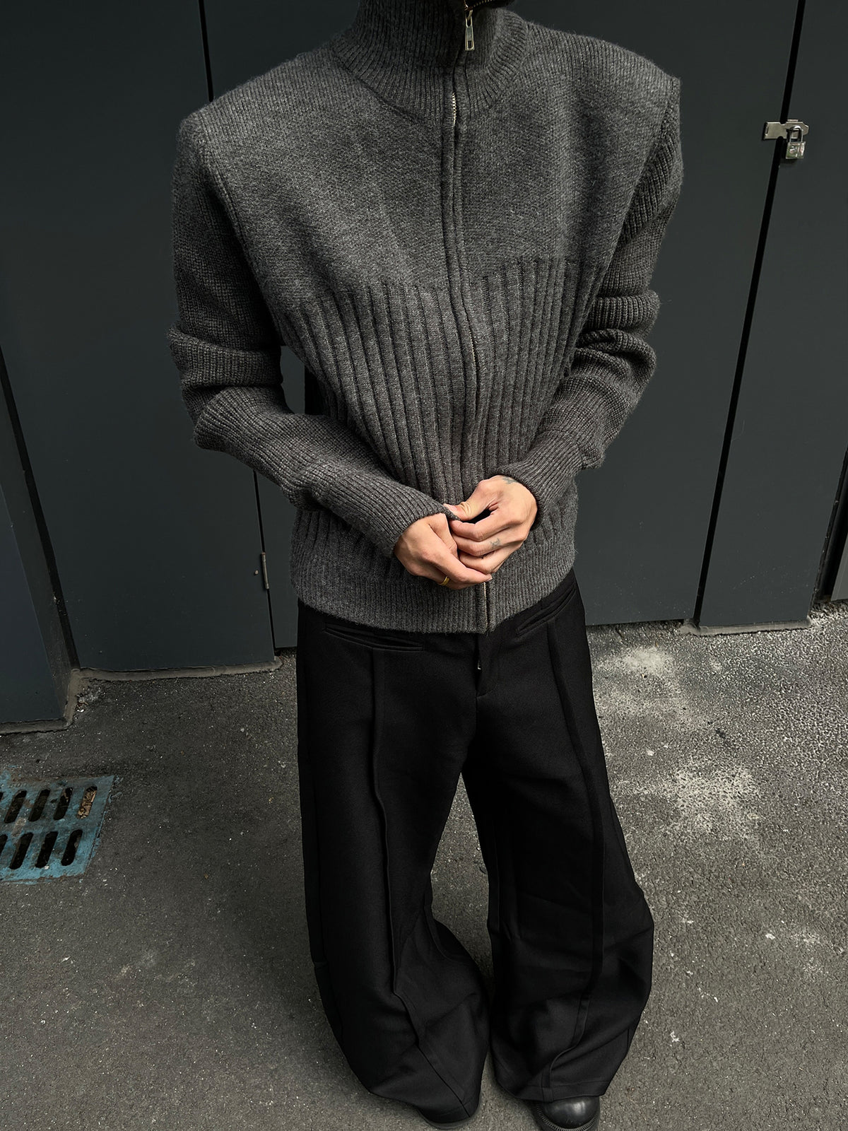 Man in the gray zip knit cardigan with hands clasped, high collar zipped up, wide black trousers
