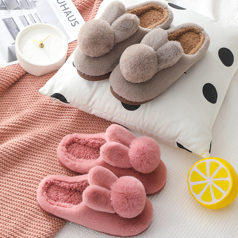 Taupe brown and burgundy plush bunny slippers with pom pom tails on a polka dot cushion beside a clock and knit throw