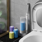 Gray toilet brush handle standing in a wall mounted holder beside a toilet, with purple, blue and yellow sponge heads