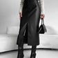 Woman in a black PU leather midi skirt with a front slit, cream sweater, black boots and a black handbag