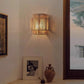 Lit hemp rope wall lamp on a cream wall beside a framed picture, a clay vase and a window