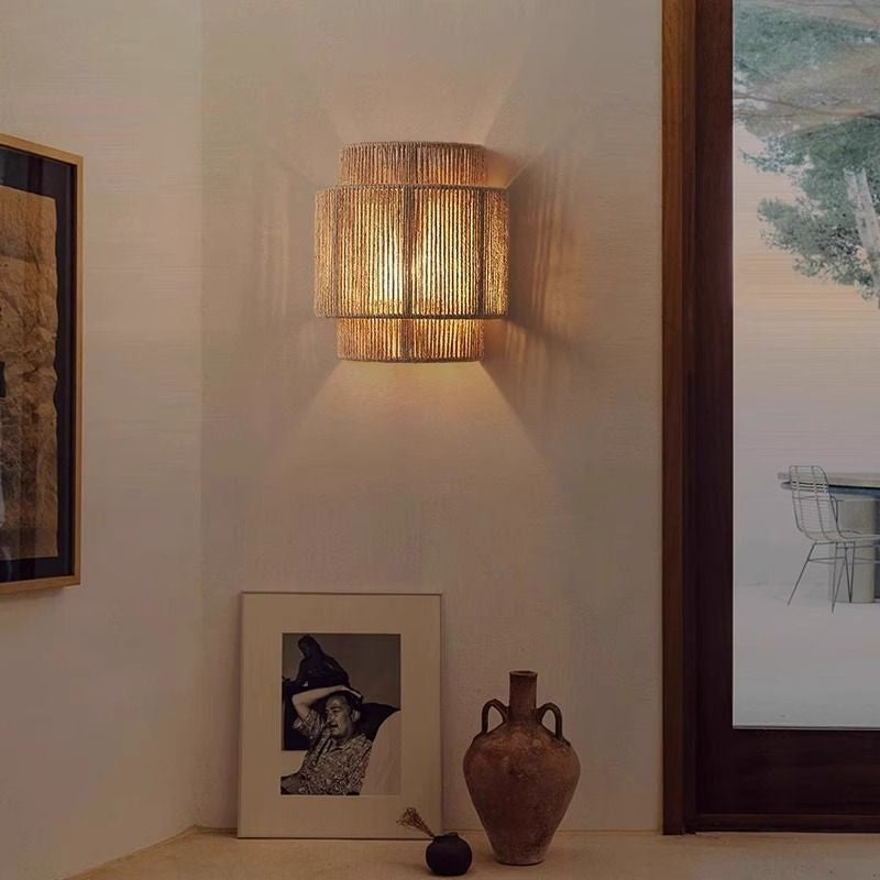 Lit hemp rope wall lamp on a cream wall beside a framed picture, a clay vase and a window