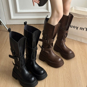 Black and brown platform buckle boots on a wood floor beside a paper shopping bag