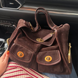 Brown suede shoulder bag with two twist lock front pockets and a buckled strap on a lap beside striped fabric