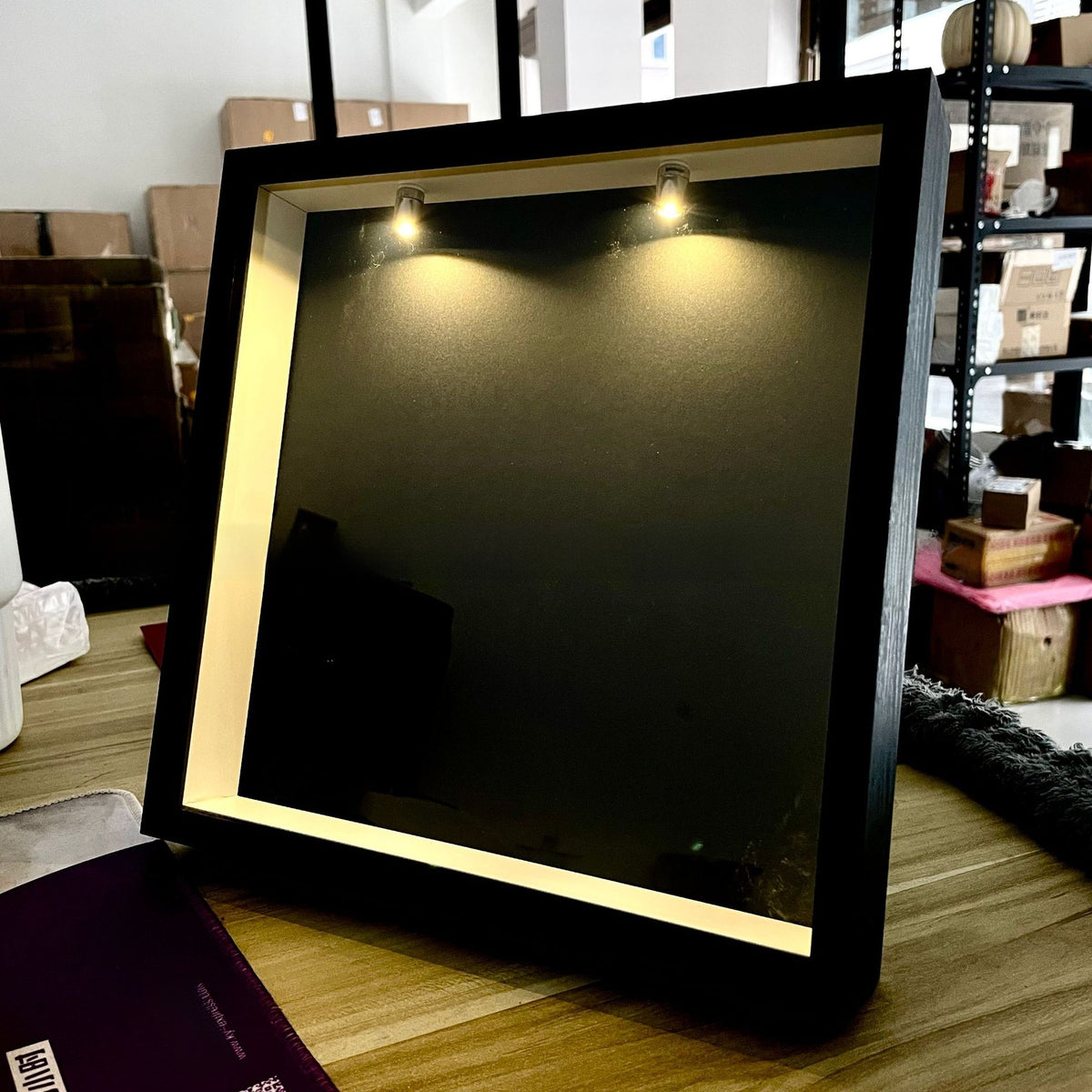 Black shadow box leaning on a table with two small spotlights lit at the top and a dark empty backdrop