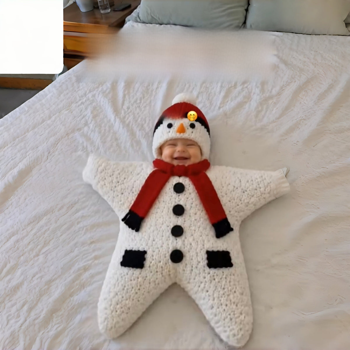 Smiling baby in a white knitted star sleeping bag with a snowman hat on a bed, snowman design not offered
