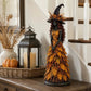 Witch statue in an orange leaf gown on a wooden console beside a lantern, candles and a pumpkin