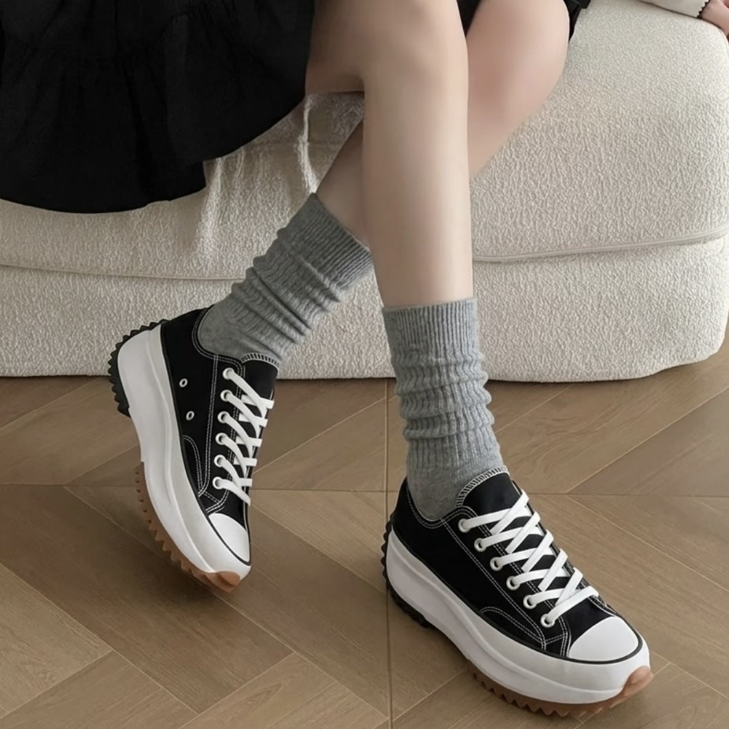 Black low-top platform canvas sneakers with white laces and a gum serrated sole worn with gray socks on a cream sofa