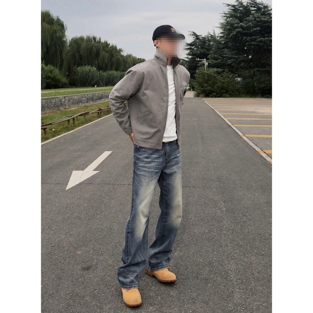 Man in a gray cotton jacket and jeans standing on a paved road, hands in the pockets