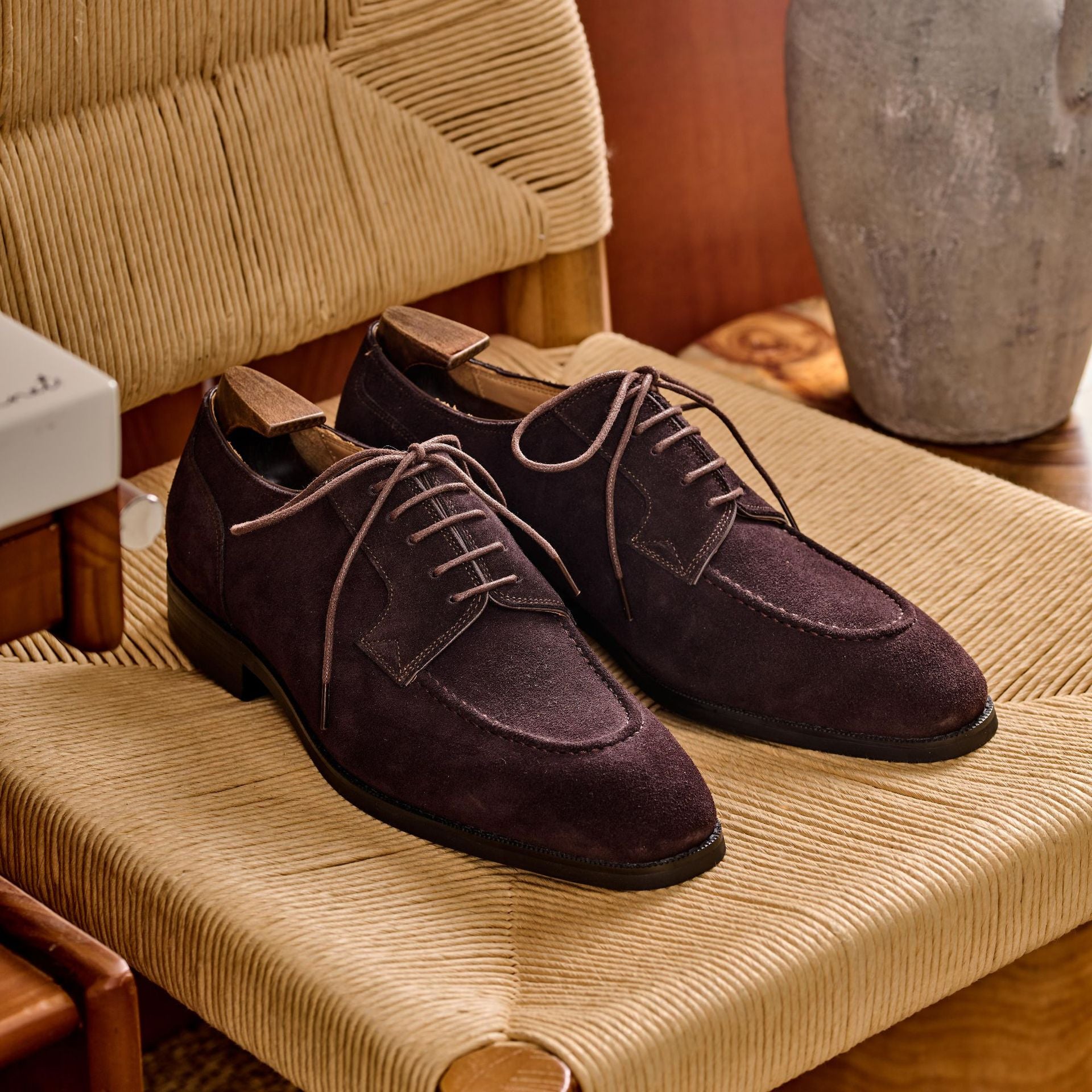 Dark brown suede derby pair on a rush chair beside a stone vase, lace panels stitched over the vamp