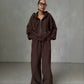 Woman wearing a brown zip-up hoodie and wide leg trousers set against a grey wall, full length view
