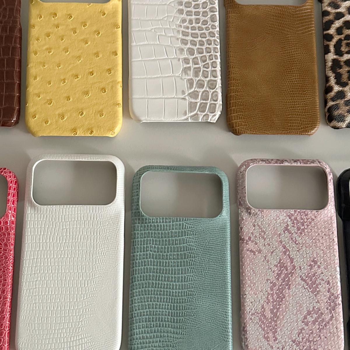 Half-cover phone cases in yellow ostrich, white crocodile, tan lizard, teal and pink snake prints on a grey surface