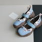 Pair of sky blue platform shoes with a wide strap, patterned panels and a brown lug sole on a grey and black surface