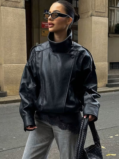 Woman in sunglasses on a city street wearing a black oversized PU leather moto jacket with a high stand collar