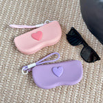 Pink and purple silicone sunglasses cases with a raised heart and wrist lanyard beside black sunglasses