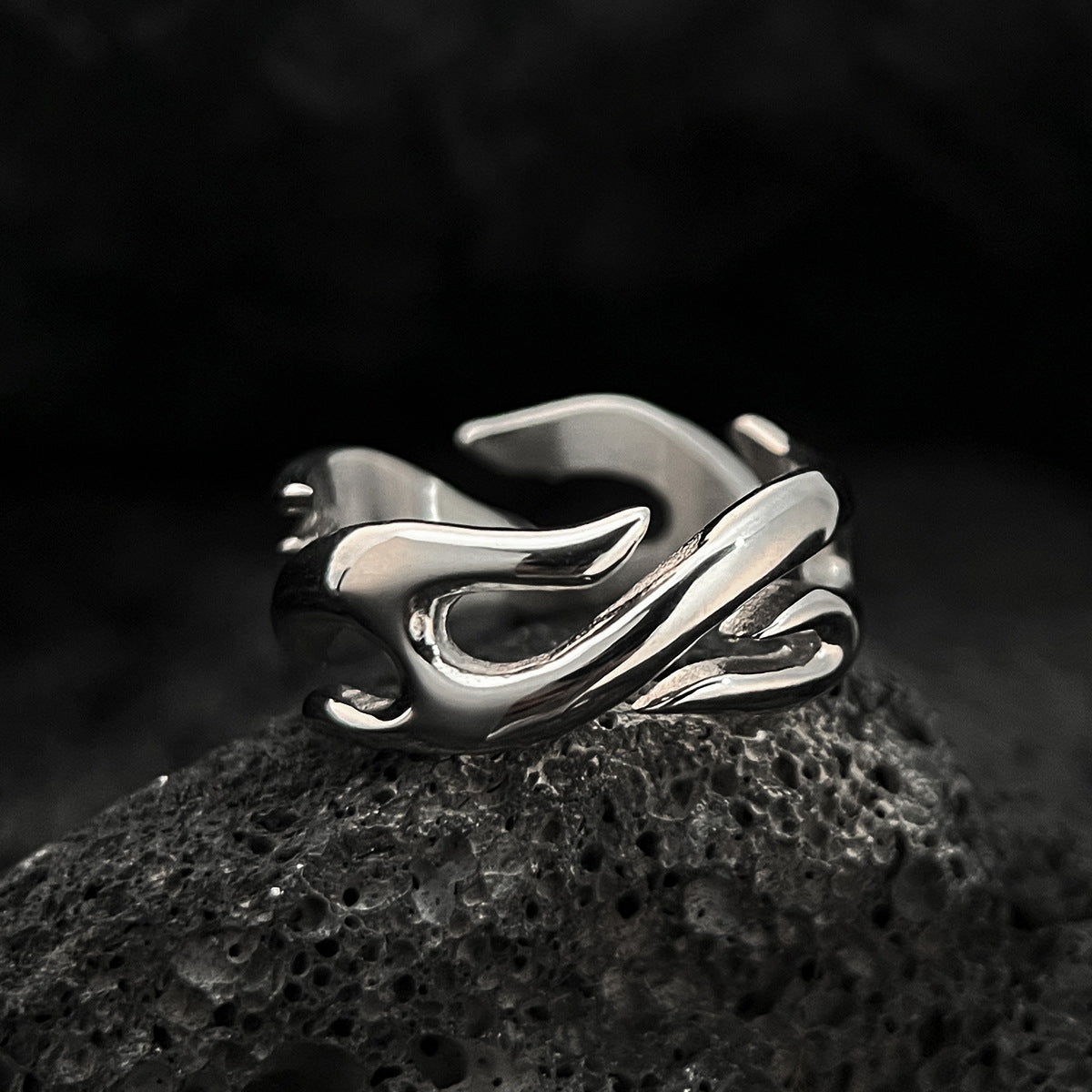 Silver tone stainless steel flame ring resting on dark porous lava rock, polished tongues catching a hard edge of light