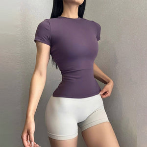 Woman in a dark purple fitted short sleeve fitness T-shirt with light gray shorts, three-quarter studio view