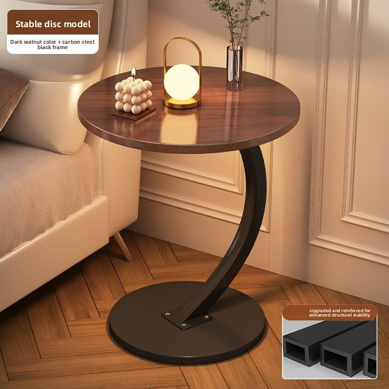Dark walnut tone round top side table with a black steel C frame and disc base beside a bed, option Dark Walnut