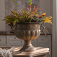 Aged bronze iron urn with scroll handles holding a bunch of ferns on a wooden table in a bright room