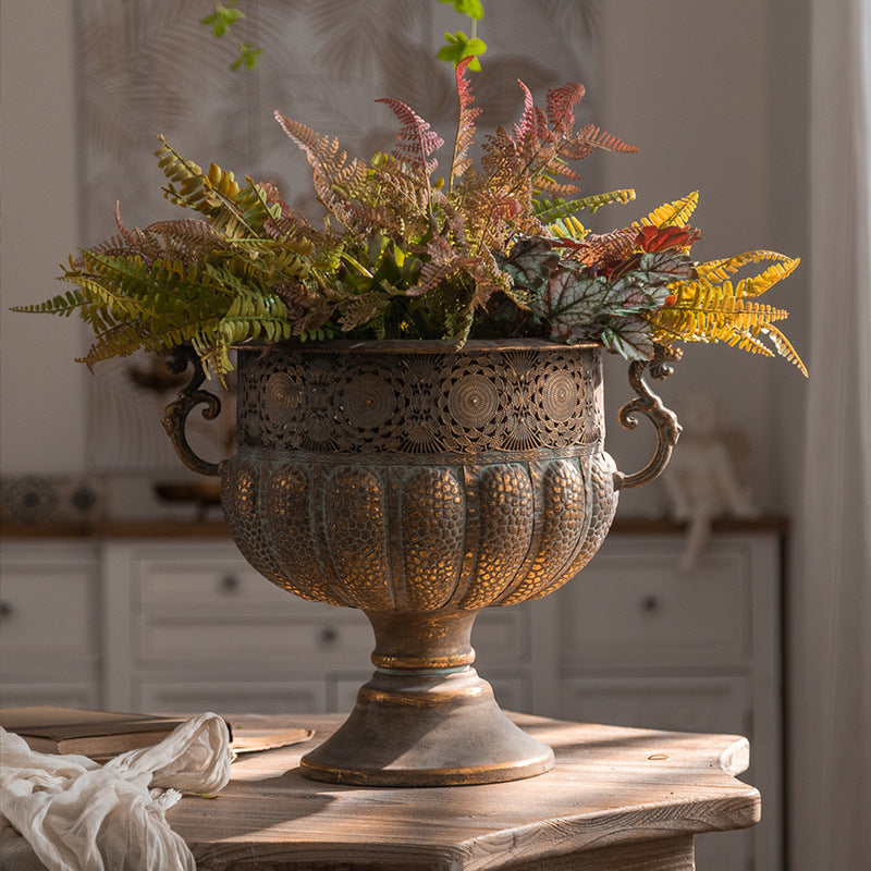 Aged bronze iron urn with scroll handles holding a bunch of ferns on a wooden table in a bright room
