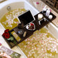 Dark brown bathtub caddy tray across a tub with a tablet, a glass of red wine, fruit, a candle and red roses