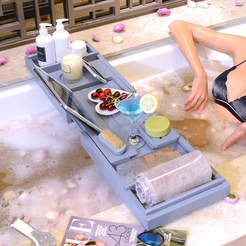 Gray bathtub caddy tray across a tub with bottles, a candle, fruit, a drink and a rolled towel, a woman bathing