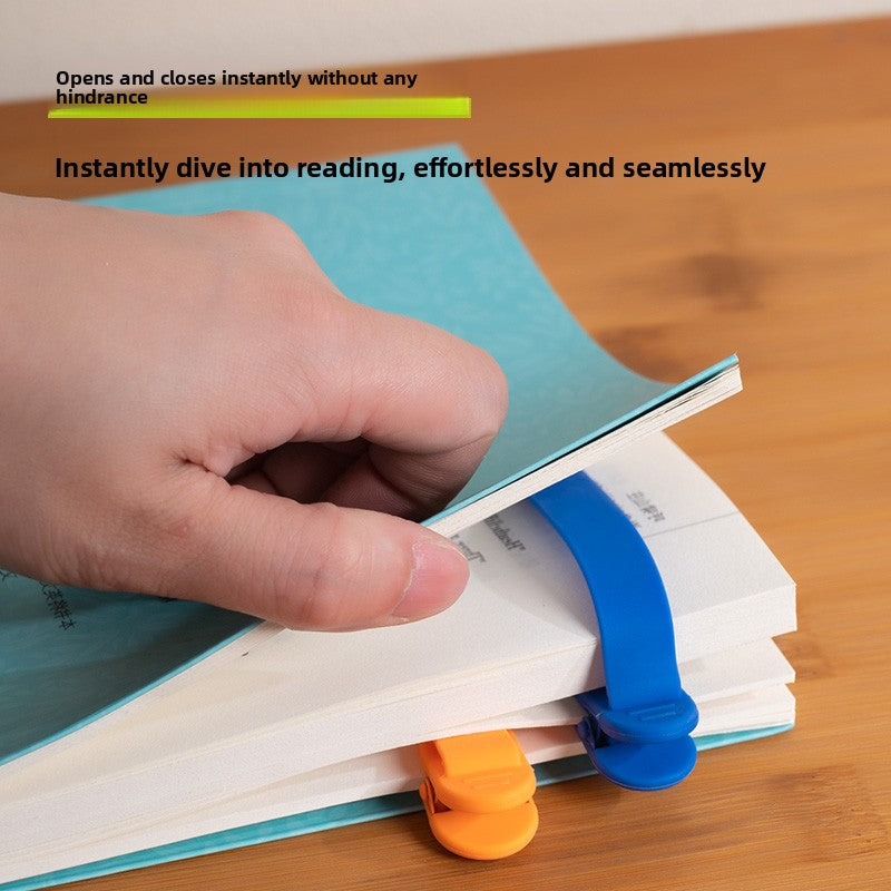 Hand slipping a blue silicone bookmark clip onto the pages of an open paperback, with an orange clip below