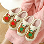 Two pairs of cream plush slippers with tufted reindeer faces with red and green noses on a pink fleece blanket