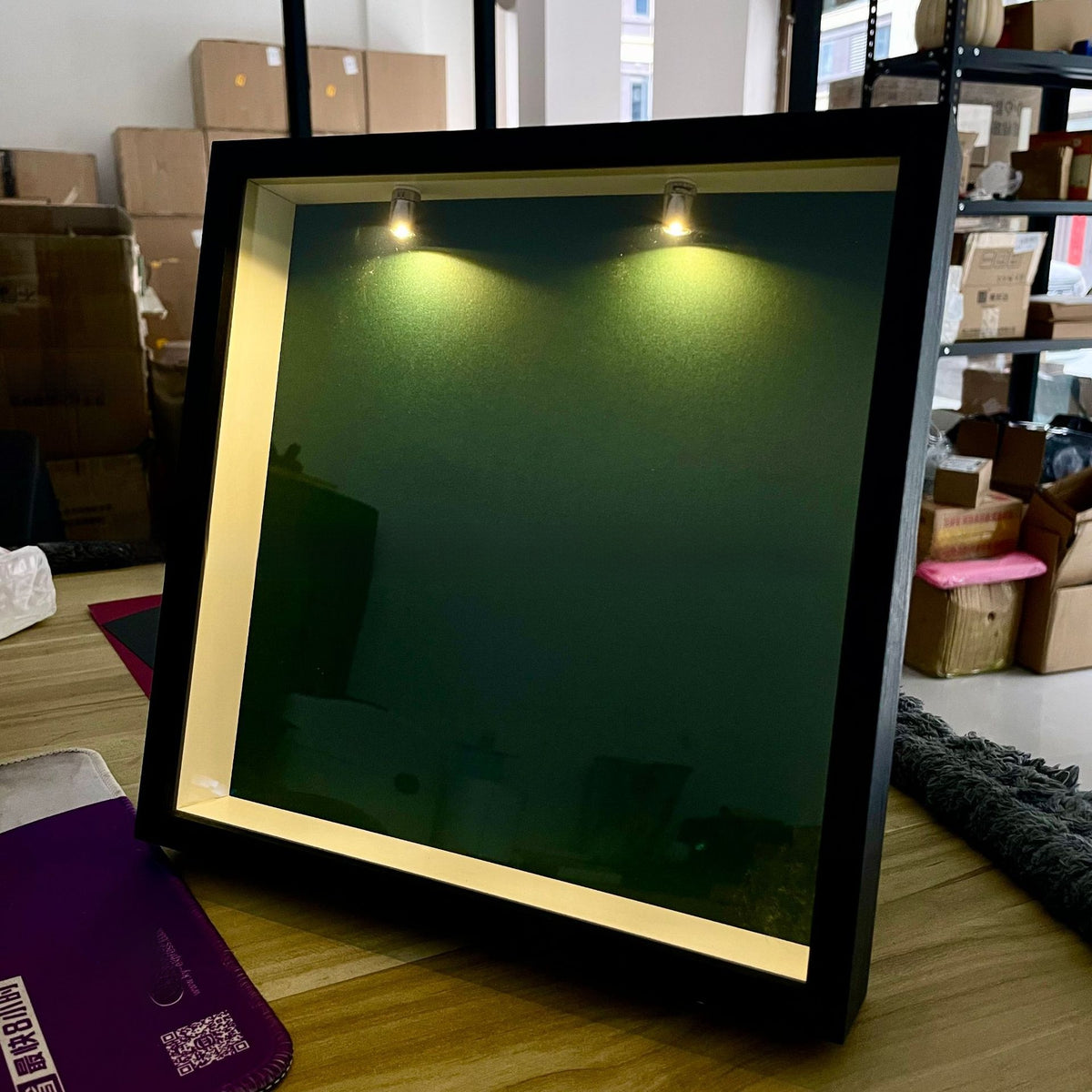 Black frame shadow box with a green backdrop and two lit spotlights at the top, photographed on a table