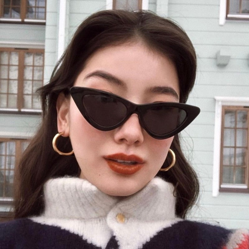 Woman wearing black triangular cat eye sunglasses with gray lenses in front of a building