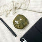 Plain olive resin frog earbud case seen from above on white cloth beside a pen and a tablet