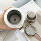 Open white flask beside its steel screw cap and the perforated tea strainer basket
