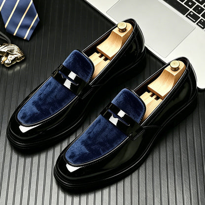 Pair of glossy black loafers with a navy velvet vamp and a penny strap on a dark desk beside a striped tie